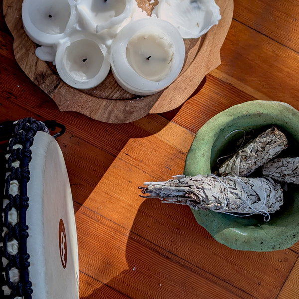 A drum, candles, and three bundles of wild sage in a green bowl, all on a pine floor.